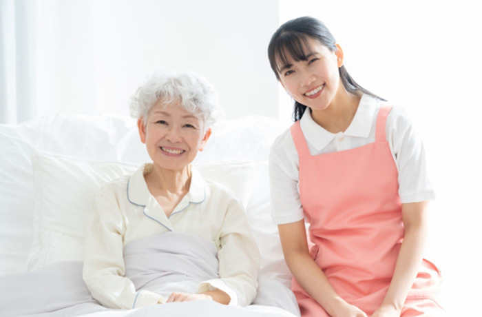 明るい部屋のベッドで笑顔でこちらを向く高齢女性と優しそうなスタッフ